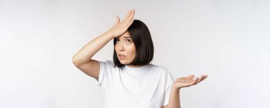 Kızgın Koreli kız facepalm, tokat alnı ve omuz silkme, pürüzsüzlükten kafası karışmış, beyaz arka planda duruyor.