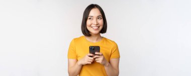 İnsanlar ve teknoloji kavramı. Gülümseyen Asyalı kız akıllı telefon kullanıyor, cep telefonuyla mesajlaşıyor, beyaz arka planda duruyor.