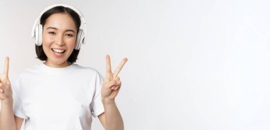 Mutlu Asyalı kadın kulaklık takıyor ve gülümsüyor, barış işareti gösteriyor, müzik dinliyor, beyaz arka plan üzerinde tişörtle duruyor.