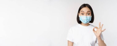 Covid-19, sağlık ve tıbbi konsept. Medikal maskeli, etkilenmiş Asyalı kadın, şaşırmış görünüyor ve tamam işareti gösteriyor, beyaz arka planda duruyor.