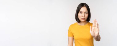 Asyalı kadın portresi ciddi ve sinirli görünüyor, dur işareti, tabu işareti, müstehcenliği yasaklıyor, beyaz arka plan üzerinde sarı tişörtle duruyor.