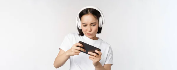 Ευτυχισμένη Ασιάτισσα με ακουστικά, κοιτάζοντας smartphone, βλέποντας βίντεο στο κινητό τηλέφωνο και χαμογελώντας, στέκεται πάνω από το λευκό φόντο — Φωτογραφία Αρχείου