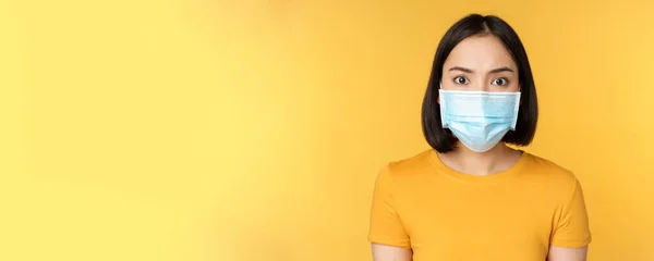 Retrato de chocado ásia mulher olhando preocupado e assustado no câmara vestindo covid-19 médico rosto máscara de pé contra amarelo fundo — Fotografia de Stock