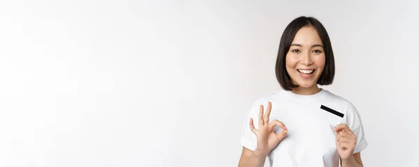 Souriant coréen fille montrant signe correct et recommandant carte de crédit de copie banque de l'espace, debout dans le t-shirt sur fond blanc — Photo