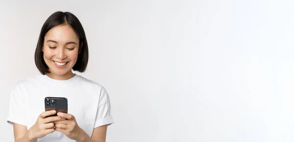 Портрет усміхненої азіатки, що використовує мобільний телефон, балачки, смс-повідомлення, стоїть у сорочці на білому тлі — стокове фото