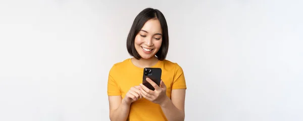 Pessoas e conceito de tecnologia. Sorrindo menina asiática usando smartphone, mensagens de texto no telefone móvel, de pé contra fundo branco — Fotografia de Stock