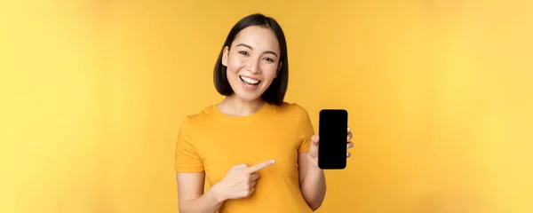 Hermosa chica asiática feliz mostrando la pantalla del teléfono móvil, aplicación en el gadget del teléfono inteligente, de pie sobre el fondo amarillo — Foto de Stock