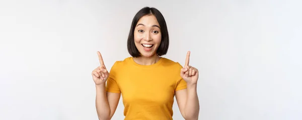 Šťastná krásná asijská dívka ukazuje reklamu, ukazuje prsty nahoru, stojící nad bílým pozadím — Stock fotografie