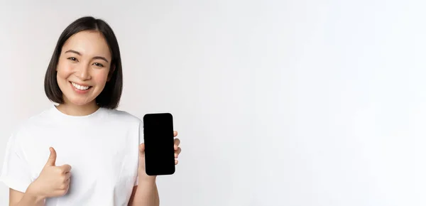 Portrait of beautiful asian woman showing thumb up and smartphone screen, smile while advertising mobile phone app, white background — Stok Foto