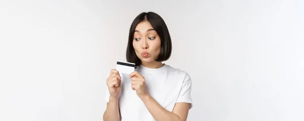 Концепція грошей та фінансів. Симпатична японська дівчина цілує свою кредитну картку, стоячи в сорочці на білому тлі — стокове фото