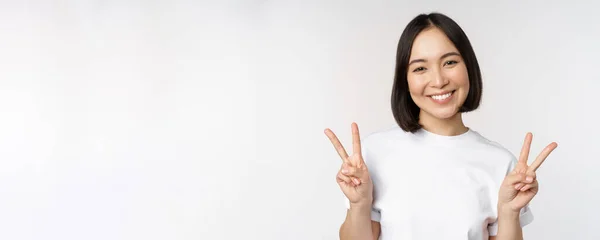 Bonito ásia menina mostrando paz, v-sign, sorrindo e olhando feliz para câmara, vestindo branco tshirt, estúdio fundo — Fotografia de Stock