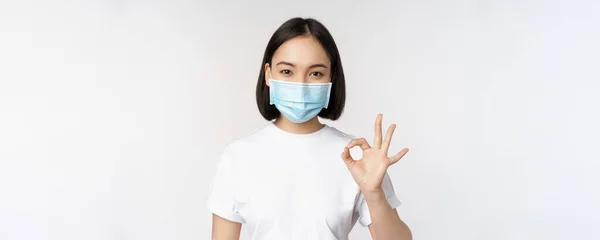 Covid-19, sağlık ve tıbbi konsept. Mutlu Asyalı kız gülümsüyor, tıbbi maske takıyor, tamam işareti gösteriyor, onaylıyor, tavsiye ediyor, beyaz arka plan — Stok fotoğraf