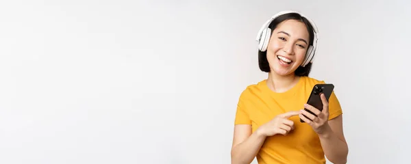 Linda chica japonesa en los auriculares, mirando el teléfono móvil y sonriendo, utilizando la aplicación de música en el teléfono inteligente, de pie sobre fondo blanco — Foto de Stock