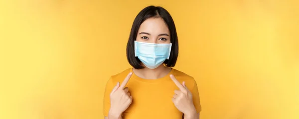 Retrato de mujer asiática sonriente con máscara facial médica, señalando su equipo de protección personal de covid-19 durante la pandemia, de pie sobre fondo amarillo — Foto de Stock