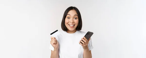 Compras online. Feliz mulher asiática usando cartão de crédito e aplicativo de smartphone, pagando no site via telefone celular, fundo branco — Fotografia de Stock