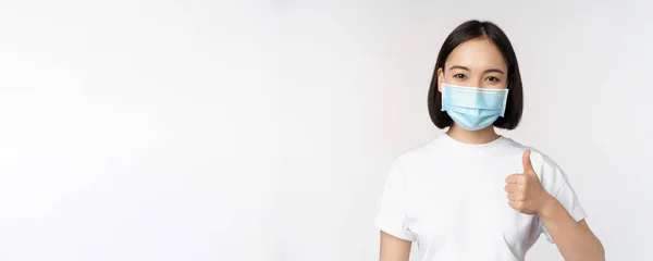 Χαμογελαστή Ασιάτισσα με ιατρική μάσκα που δείχνει τους αντίχειρες της, εγκρίνει κάτι καλό, επαίνους και κομπλιμέντα παρέα, στέκεται πάνω από το λευκό φόντο — Φωτογραφία Αρχείου