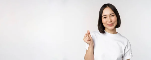Hermosa mujer asiática sonriendo, mostrando el gesto de los corazones de los dedos, usando camiseta, de pie sobre fondo blanco — Foto de Stock