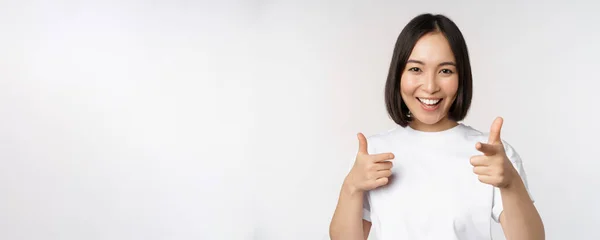 Image d'une fille asiatique souriante pointant du doigt la caméra, choisissant, vous invitant, félicitant, debout en t-shirt sur fond blanc — Photo