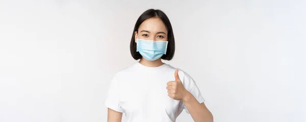 Uśmiechnięta Azjatka w masce medycznej pokazująca kciuki do góry, zatwierdzająca coś dobrego, chwaląca i komplementująca towarzystwo, stojąca na białym tle — Zdjęcie stockowe