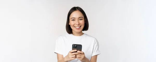 Porträtt av leende asiatisk kvinna med hjälp av mobiltelefon, chatta, sms: a meddelande, står i t-shirt över vit bakgrund — Stockfoto
