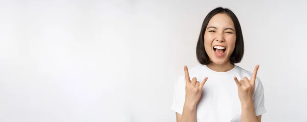 Напористая азиатка кричит, наслаждается концертом или фестивалем, показывает рок на, хеви-метал знак, весело, стоя на белом фоне — стоковое фото