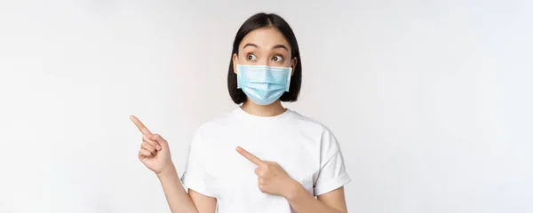 Ung koreansk kvinna i medicinsk ansiktsmask pekar finger vänster och tittar på logotyp, visar reklam eller banner, står över vit bakgrund — Stockfoto