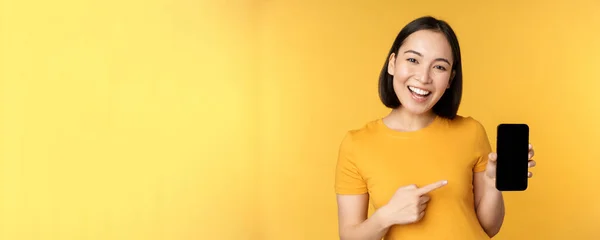 Bella ragazza asiatica felice che mostra lo schermo del telefono cellulare, applicazione su gadget smartphone, in piedi su sfondo giallo — Foto Stock