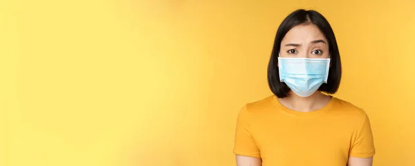 Portrait of skeptical and confused asian woman in medical face mask, raising eyebrow doubtful, standing over yellow background — Stock Photo, Image