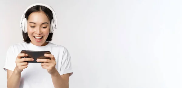 Heureuse femme asiatique dans les écouteurs, regardant smartphone, regardant la vidéo sur téléphone mobile et souriant, debout sur fond blanc — Photo