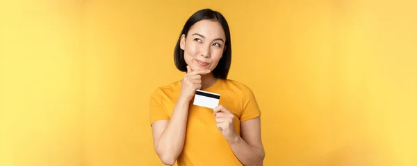 Pensando mujer asiática sosteniendo tarjeta de crédito y sonriendo, planeando comprar smth, de pie reflexivo sobre fondo amarillo — Foto de Stock