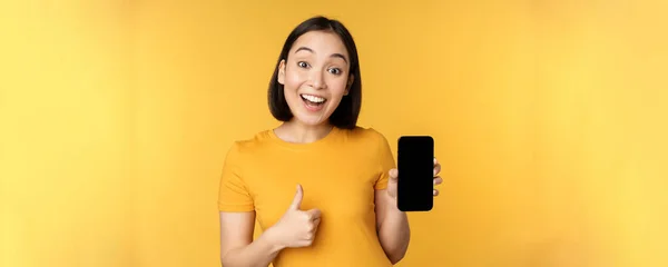 Spännande asiatisk kvinna som visar mobiltelefon skärm, tummen upp, som smth bra, rekommendera smartphone ansökan, står över gul bakgrund — Stockfoto