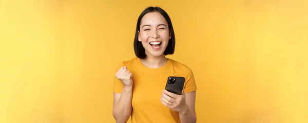 Joyful asian woman celebrating, holding mobile phone, winning, achieve goal on smartphone, standing over yellow background — Stock Photo, Image