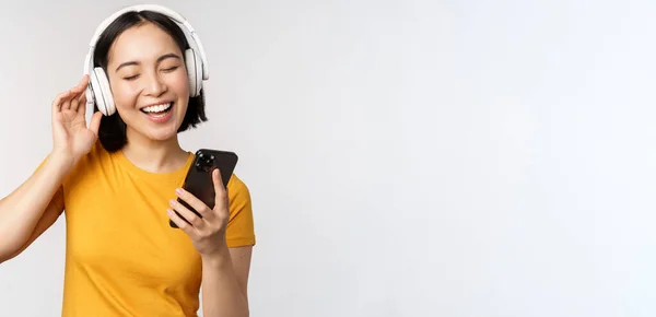 Linda chica japonesa en los auriculares, mirando el teléfono móvil y sonriendo, utilizando la aplicación de música en el teléfono inteligente, de pie sobre fondo blanco — Foto de Stock