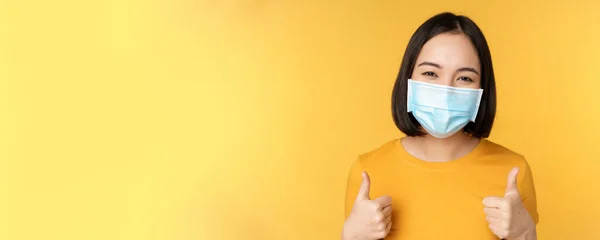 Mulher coreana alegre em máscara facial médica, apoiar as pessoas durante a pandemia, usar equipamentos de proteção individual de covid-19, mostrando polegares para cima na aprovação, fundo amarelo — Fotografia de Stock