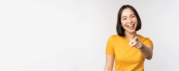 Feliz hermosa mujer asiática riendo, señalando con el dedo a la cámara y riendo, sonriendo despreocupado, de pie en camiseta amarilla sobre fondo blanco — Foto de Stock