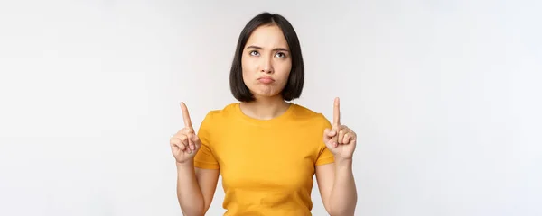 Enttäuschte asiatische Frau schaut, zeigt mit erhobenen Fingern und wütendem launischen Gesichtsausdruck in gelbem T-Shirt vor weißem Hintergrund — Stockfoto