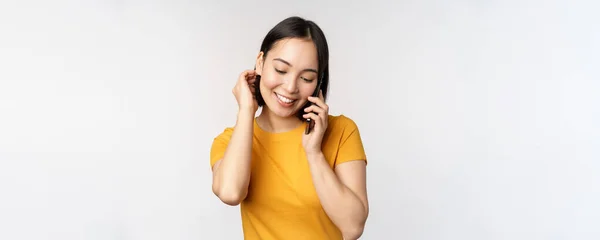 낭만적 이고 귀여운 동양 소녀는 전화를 받고, 휴대 전화로 이야기하고, 웃으며, 하얀 배경에 노란 티셔츠를 입고 서 있다 — 스톡 사진
