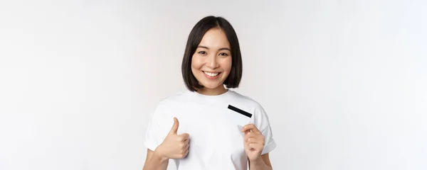 Portré gyönyörű fiatal modern ázsiai nő, mutatja a hitelkártya és hüvelykujj fel, ajánló érintésmentes fizetés, áll a fehér háttér — Stock Fotó