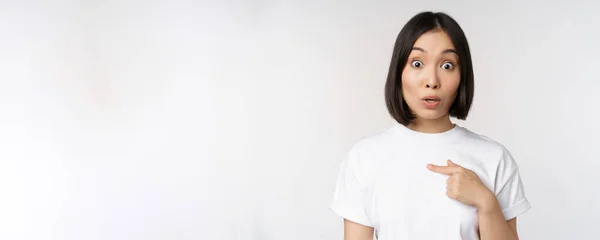 Retrato de surpreendido asiático menina estudante, apontando dedo esquerdo, levantando sobrancelhas espantado, impressionado com grande negócio, oferecer à frente, de pé sobre fundo branco — Fotografia de Stock