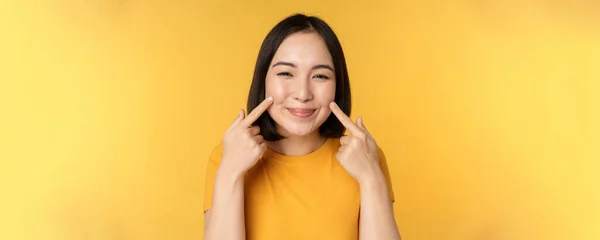 De cerca retrato de linda chica asiática mostrando sus hoyuelos y sonriente coqueta en la cámara, de pie sobre el fondo amarillo —  Fotos de Stock