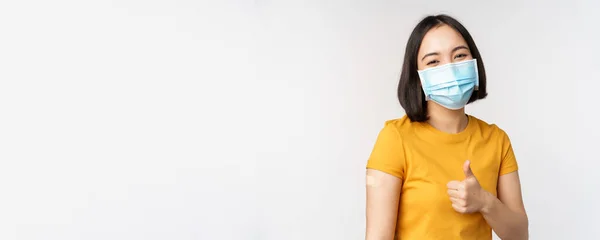 Covid-19, vaccination and healthcare concept. Portrait of cute asian girl in medical mask, has band aid on shoulder after coronavirus vaccine, standing over white background — Stock Photo, Image