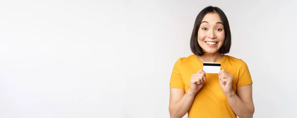 Retrato de hermosa chica coreana sosteniendo tarjeta de crédito, recomendando servicio bancario, de pie en camiseta amarilla sobre fondo blanco —  Fotos de Stock