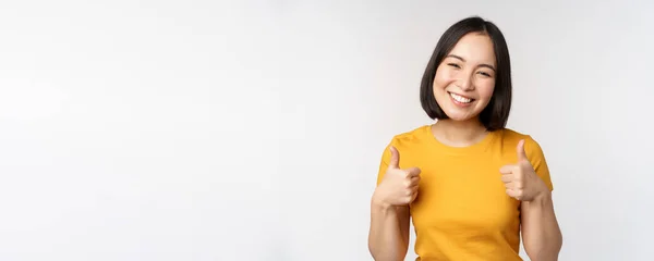 Magnifique modèle féminin asiatique souriant, montrant les pouces levés et regardant heureux, recommandant, exprimant une rétroaction positive, debout sur fond blanc — Photo