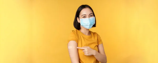 Vaccinatie en covid-19 pandemie concept. Glimlachende Aziatische vrouw met medisch gezichtsmasker, die haar schouder laat zien met pleister na vaccinatie met coronavirus, gele achtergrond — Stockfoto