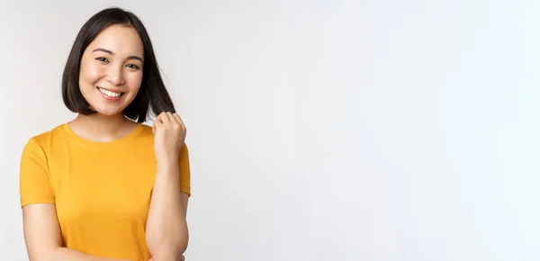Linda menina asiática romântica, sorrindo e brincando com o cabelo, olhando feliz para a câmera, de pé em camiseta amarela sobre fundo branco — Fotografia de Stock