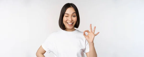Va tutto bene. Sorridente giovane donna asiatica assicurando, mostrando ok segno con volto soddisfatto, in piedi su sfondo bianco — Foto Stock