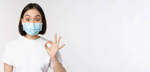 Covid-19, santé et concept médical. Femme asiatique impressionnée dans le masque médical, regardant étonné et montrant ok, ok signe, debout sur fond blanc — Photo
