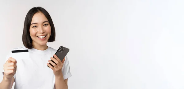 Le asiatisk flicka med mobiltelefon, visar kreditkort, begreppet kontaktlös betalning, online shopping, står över vit bakgrund — Stockfoto