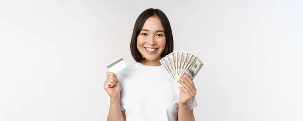 Portrait of asian woman smiling, holding credit card and money cash, dollars, standing in tshirt over white background — Stok Foto