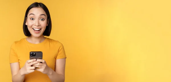 Felice ragazza asiatica sorridente, in piedi con il telefono cellulare nero, in piedi contro sfondo giallo — Foto Stock